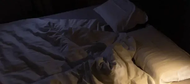 Dimly lit unmade bed with rumpled sheets and soft shadows, evoking absence and quiet emotional strain.