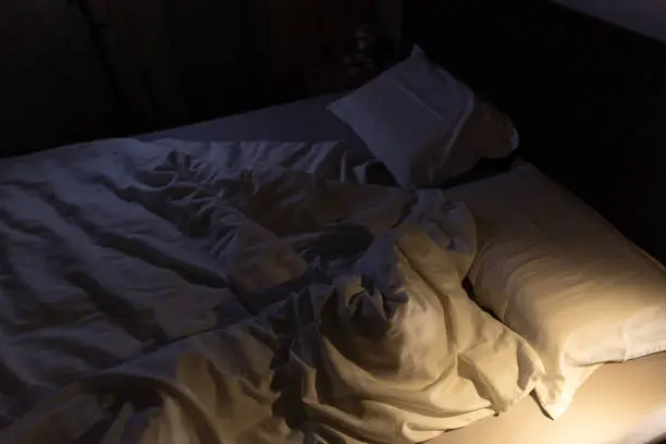 Dimly lit unmade bed with rumpled sheets and soft shadows, evoking absence and quiet emotional strain.