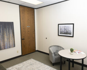 Quiet therapy office corner with a wooden door labeled ‘Please Do Not Disturb Session in Progress,’ a gray chair beside a round table holding a small plant and magazines, and framed artwork on cream walls. The space feels calm, private, and professional.